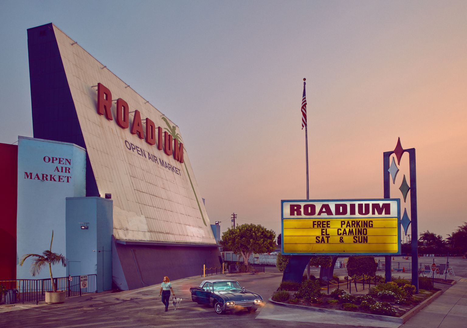 Photograph of a vintage car parked outside a mid-century modernist roadside market building at dusk.